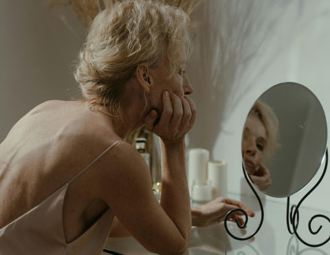 Mujer de piel madura frente al espejo realizando cuidado facial con el contorno de ojos antienvejecimiento para reducir bolsas y mejorar el drenaje linfático. 